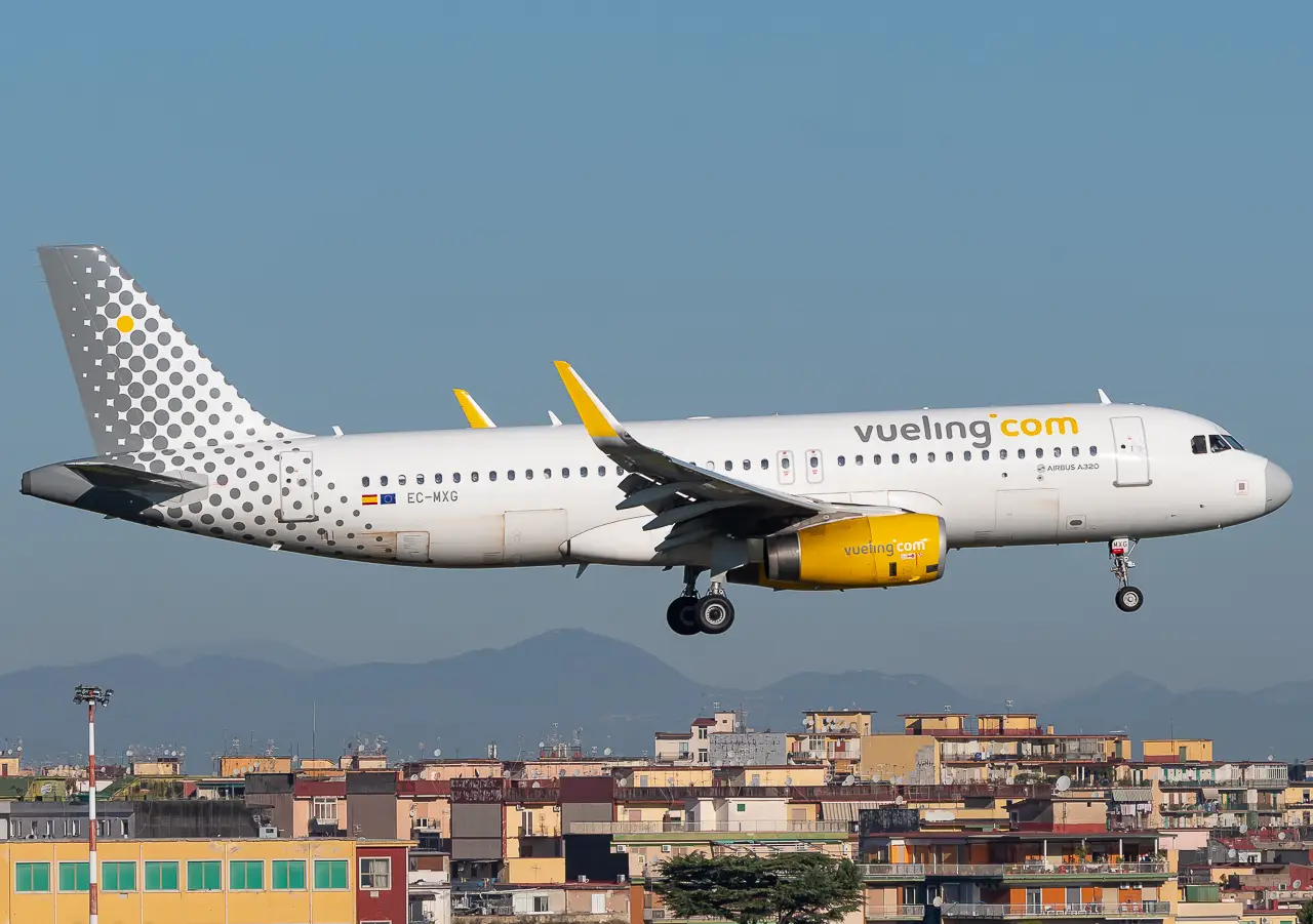 Sarı motor kapaklı ve noktalı kuyruk tasarımına sahip Vueling Airbus A320 uçağı, arka planda şehir manzarası ve uzaktaki dağlar eşliğinde inişe hazırlanıyor.