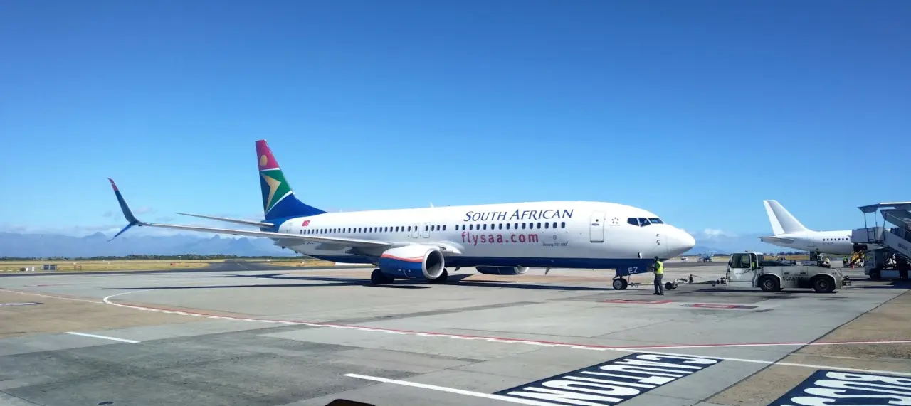 Güney Afrika Havayolları’na ait bir Boeing 737-800 uçağı, apron üzerinde kalkışa hazırlanıyor, arka planda berrak mavi bir gökyüzü ve dağ manzarası görünüyor.