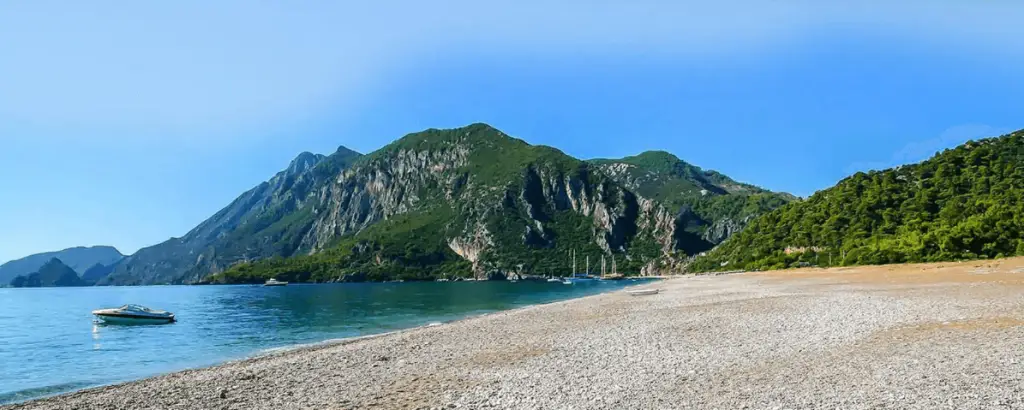 Çıralı plajı ve arka planda yemyeşil dağ manzarası. Sahil boyunca taşlık alan ve sakin bir deniz görülüyor.
