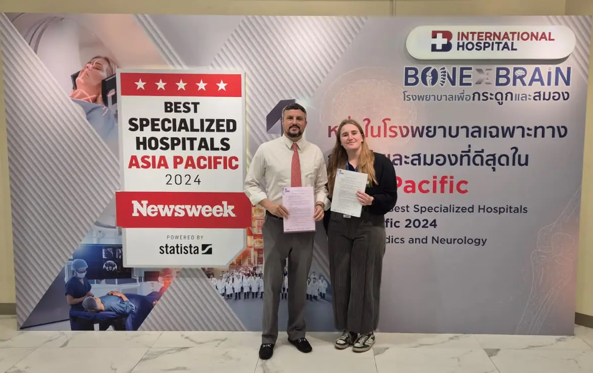 Two individuals holding signed documents stand in front of a backdrop displaying “Best Specialized Hospitals Asia Pacific 2024,” awarded by Newsweek, at an international hospital specializing in orthopedics and neurology.