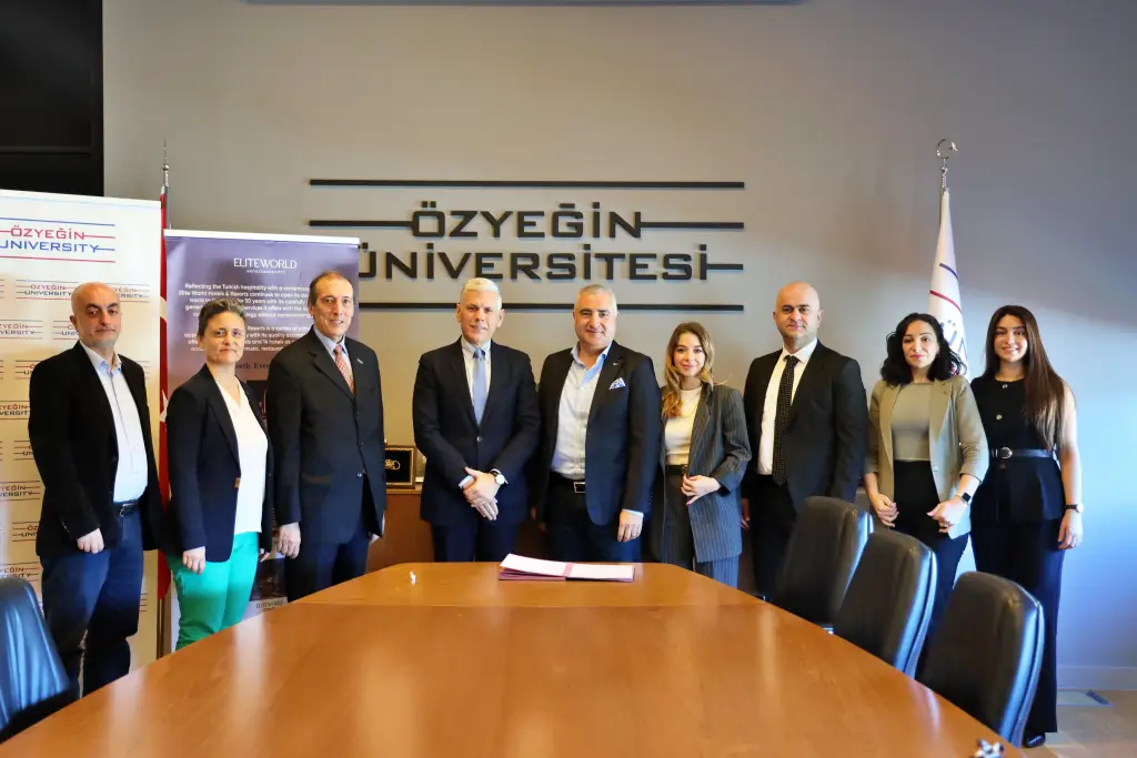 Özyeğin Üniversitesi toplantı odasında, Elite World Hotels & Resorts ve Özyeğin Üniversitesi yetkilileri, turizmde kariyer gelişimi için iş birliği protokolü imza töreninde bir araya geliyor. Masanın üzerinde imzalanmış belgeler bulunuyor, arka planda üniversitenin logosu görülüyor.