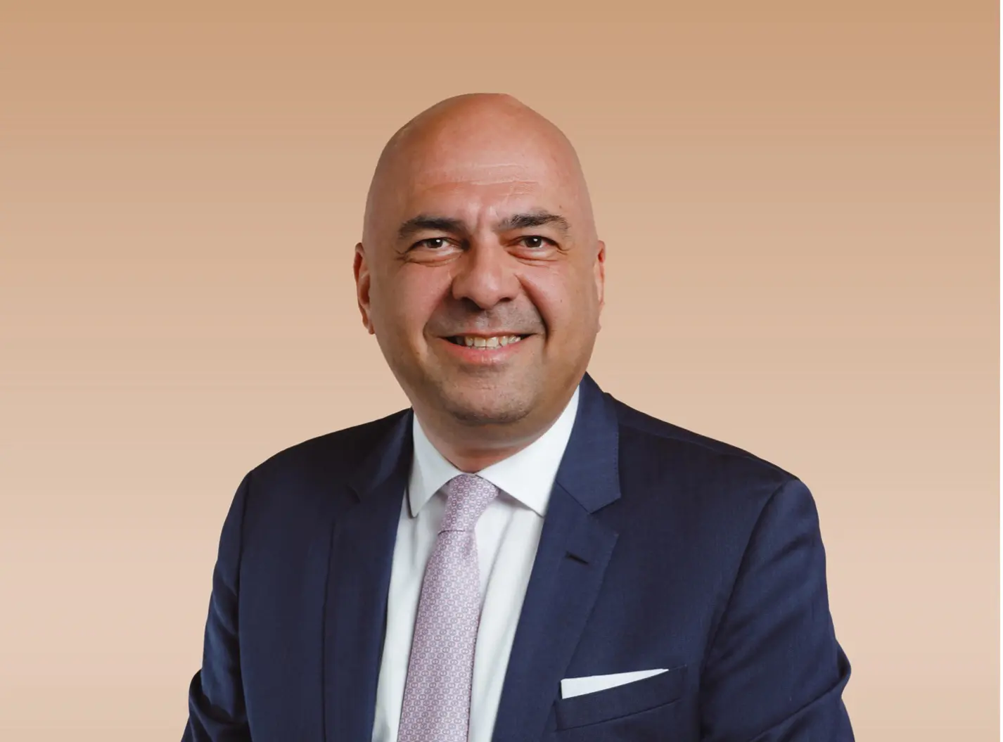 Portrait of Avsar Koc in a dark suit and tie, smiling in front of a plain beige background.