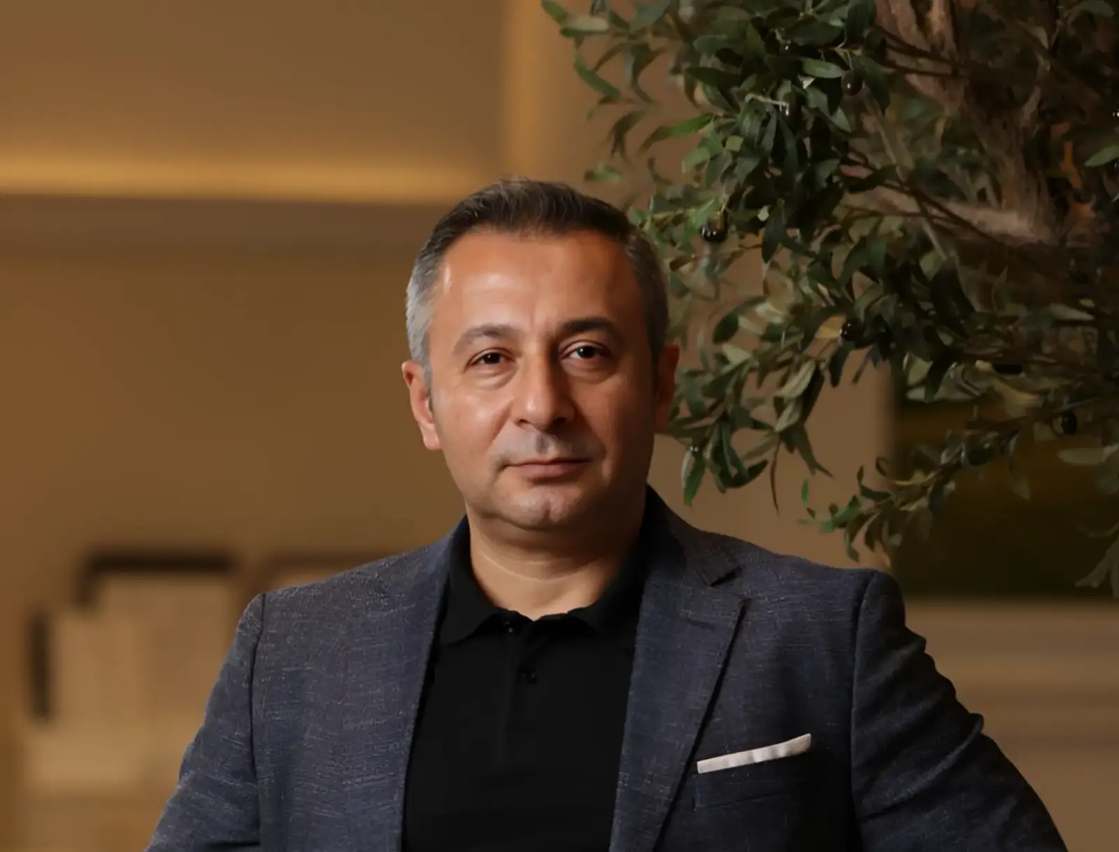 Recep Pamukcu, Ajman Hotel Resort Director, posing in a formal jacket beside an indoor olive tree