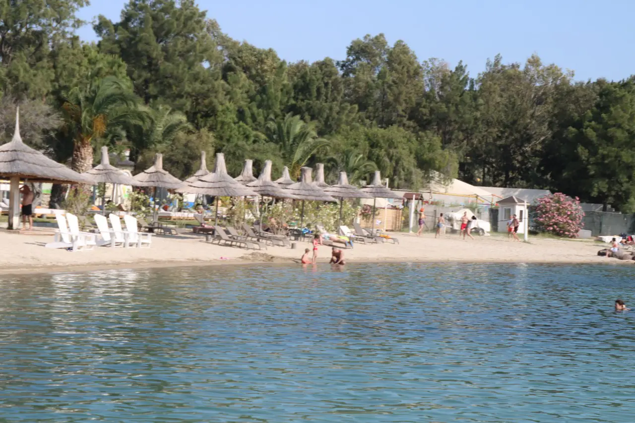 Gölgelikli şezlongların yer aldığı sakin bir Ege plajında denize giren tatilciler.