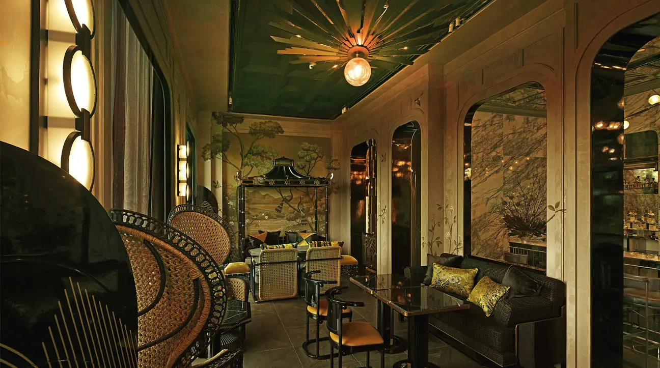 Interior of Stella bar at Capella Bangkok, featuring opulent Asian-inspired decor with hand-painted wall murals, peacock chairs, and a green coffered ceiling with a glowing central light fixture.