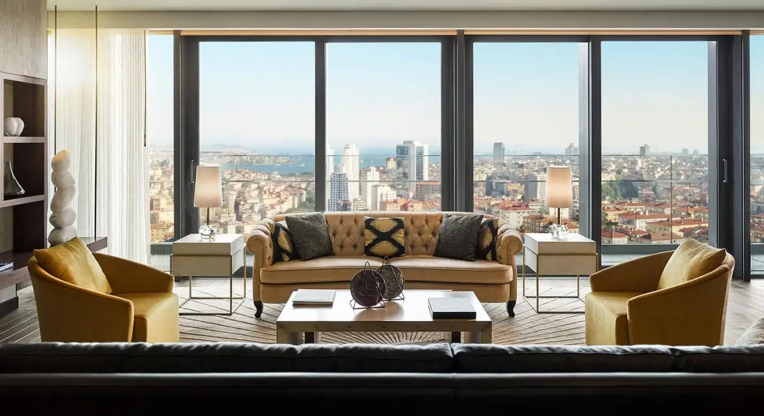 Fairmont Quasar İstanbul otelinin lüks süit odasında modern dekorasyonlu oturma alanı ve geniş pencerelerden görünen Boğaz ve şehir manzarası.