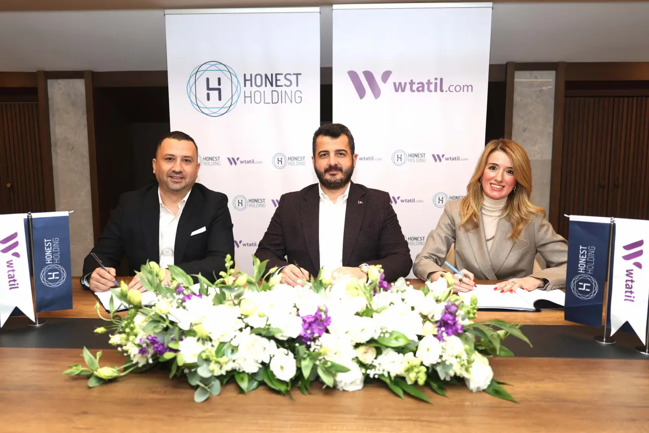 Honest Holding ve Wtatil yöneticilerinin, logolar önünde düzenlenen imza töreninde masada sözleşme imzalarken çekilmiş fotoğrafı.