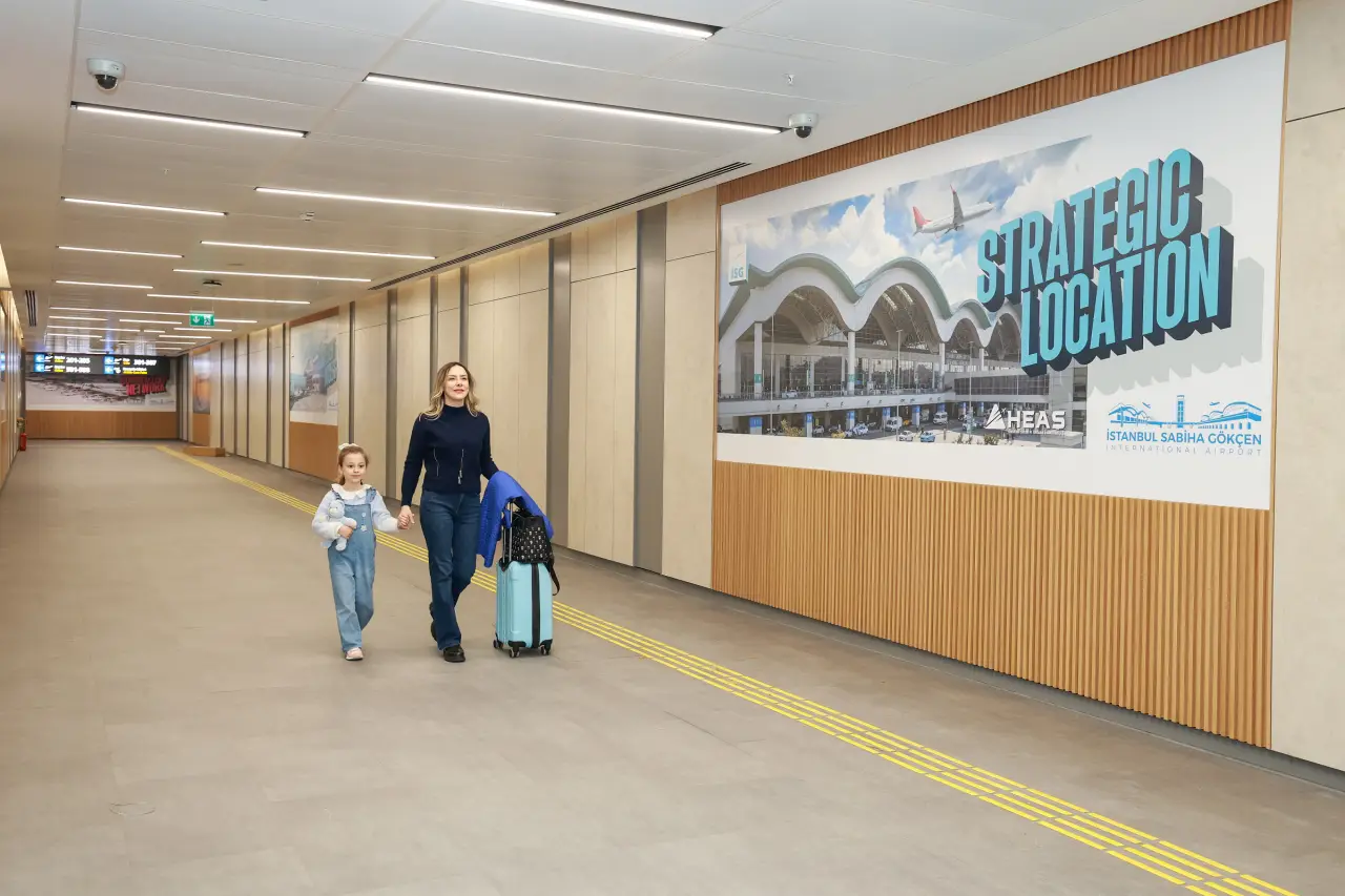 Sabiha Gökçen Havalimanı terminalinde valiziyle yürüyen anne ve çocuk, duvarda “Strategic Location” reklam panosu görülüyor.