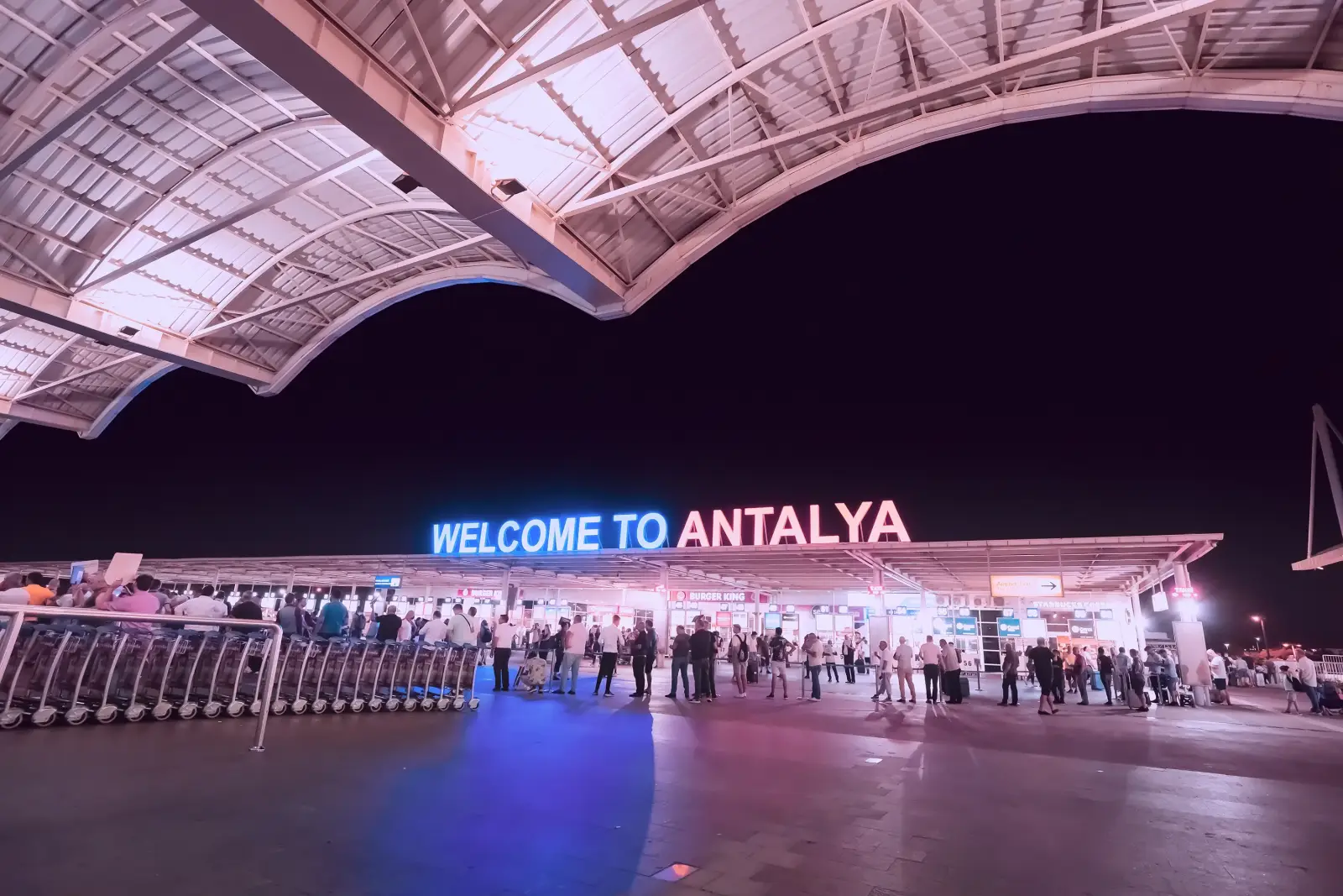 Antalya Havalimanı girişinde gece çekimi, “Welcome to Antalya” tabelası ve yolcular