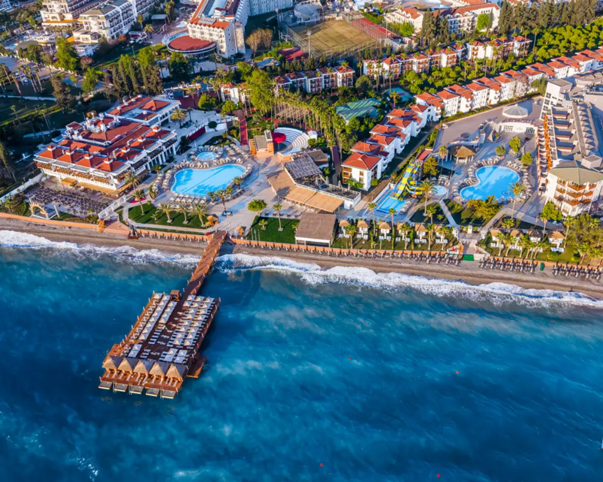 Kemer sahilinde denize sıfır konumdaki Corendon Hydros Club otelinin havadan çekilmiş fotoğrafı; ön planda ahşap iskele ve turkuaz deniz, geride havuzlar, yeşil bahçeler ve otel binaları görülüyor.