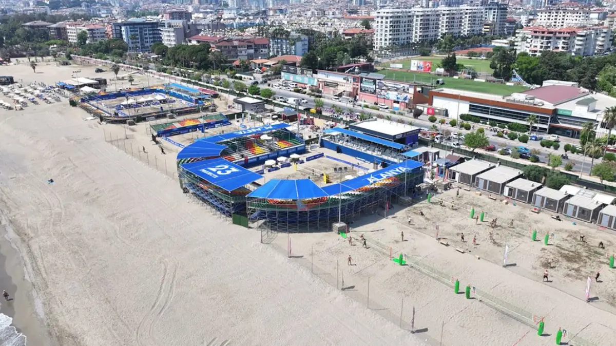 Alanya sahilinde kurulu plaj sporları merkezinin havadan görünümü: mavi tribünlü ana stadyum, çevresindeki antrenman sahalarında oynayan plaj voleybolcuları, arka planda şehir ve yeşil alanlar
