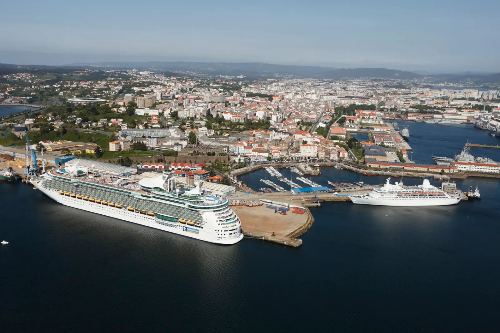 İspanya'nın kuzeyindeki Ferrol Kruvaziyer Limanı'nın havadan çekilmiş geniş açılı görünümü. Rıhtıma yanaşmış biri büyük, diğeri daha küçük iki beyaz kruvaziyer gemisi, arkalarında kentin kırmızı çatılı binaları, marina ve tekneler ile uzakta yeşil tepeler yer alıyor.