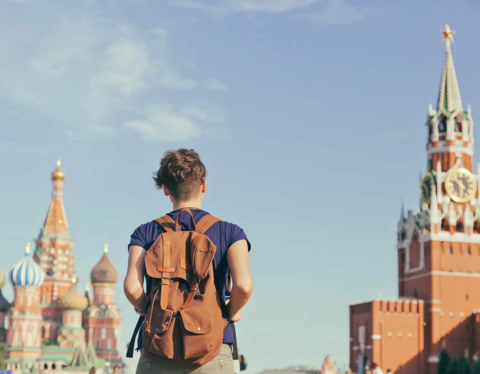 Sırt çantalı bir turist, Moskova'daki Kızıl Meydan'da Aziz Vasili Katedrali'ne ve Kremlin'in Spasskaya Kulesi'ne bakarken arkadan görüntülenmiş; mavi gökyüzü ve tarihi yapılar arka planda yer alıyor.