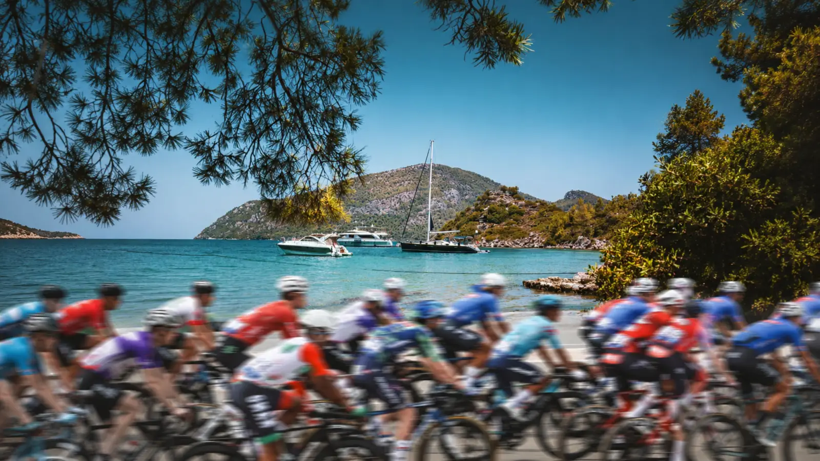 L'Étape Marmaris by Tour de France etkinliğinde turkuaz bir koy, tekneler ve çam ağaçları eşliğinde kıyı yolunda yarışan renkli formalı bisikletçi grubu