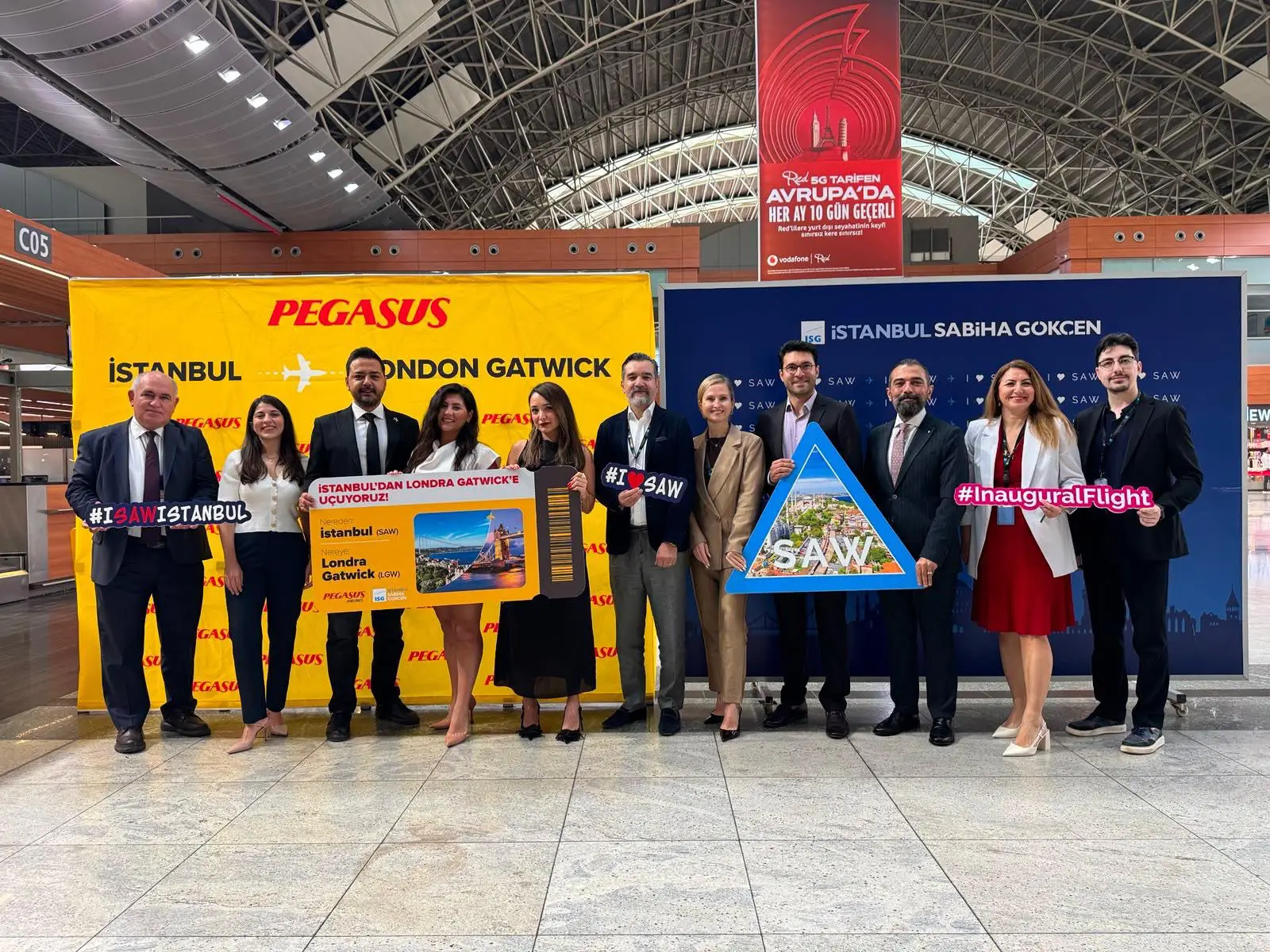 Sabiha Gökçen Havalimanı terminalinde düzenlenen tören; Pegasus Hava Yolları ve İstanbul Sabiha Gökçen yetkilileri, sarı Pegasus ve mavi ISG logolu pankartların önünde, "Istanbul'dan Londra Gatwick'e Uçuyoruz" yazılı büyük bilet görseli ve SAW logolu pankartlarla ilk seferi kutluyor.