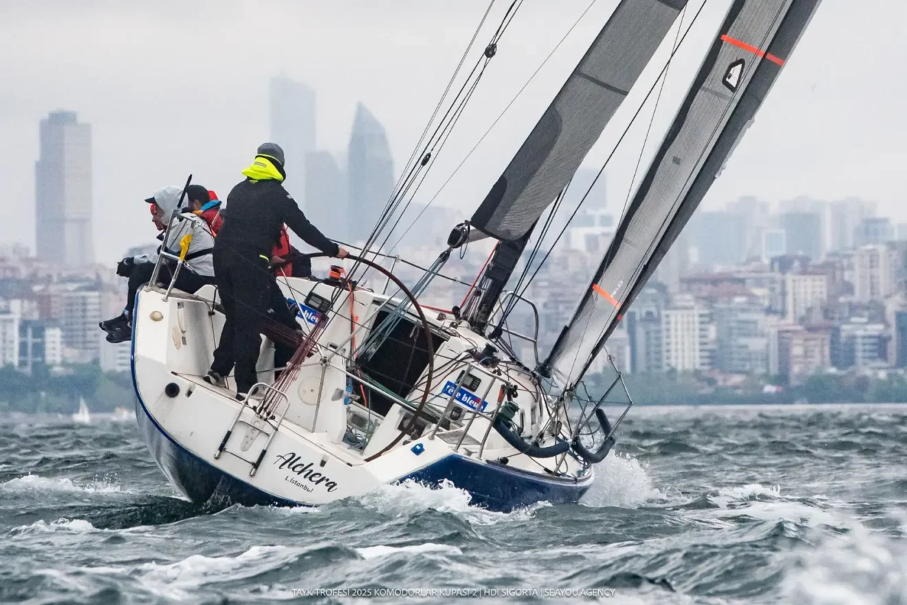 TAYK Trofesi 2025 Komodorlar Kupası-2 HDI Sigorta Kupası yarışından bir kare: "Alchera" isimli yelkenli, dalgalı suda yan yatarak seyrederken güvertede siyah yağmurluk giyen mürettebat manevra yapıyor; arka planda sisli İstanbul silueti görünüyor