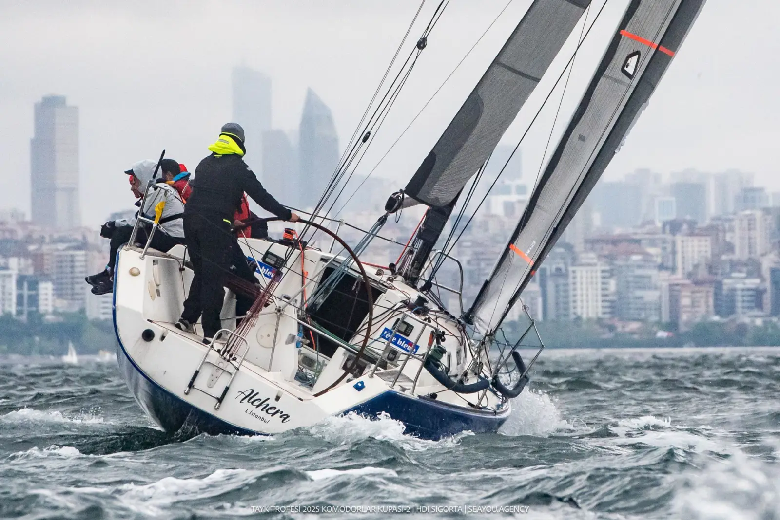 TAYK Trofesi 2025 Komodorlar Kupası-2 HDI Sigorta Kupası yarışından bir kare: "Alchera" isimli yelkenli, dalgalı suda yan yatarak seyrederken güvertede siyah yağmurluk giyen mürettebat manevra yapıyor; arka planda sisli İstanbul silueti görünüyor