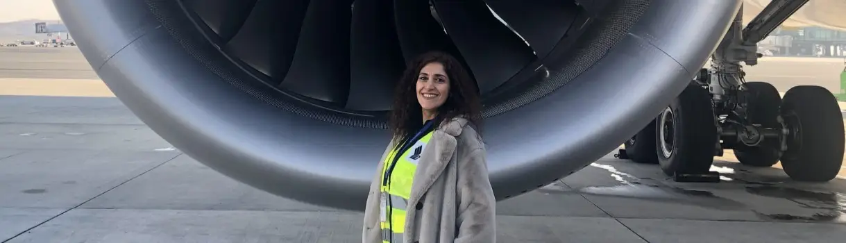 Travelport Türkiye'nin yeni Ülke Müdürü Mehtap Topcu, sarı reflektörlü yeleğiyle bir uçağın motoru önünde pist alanında gülümserken