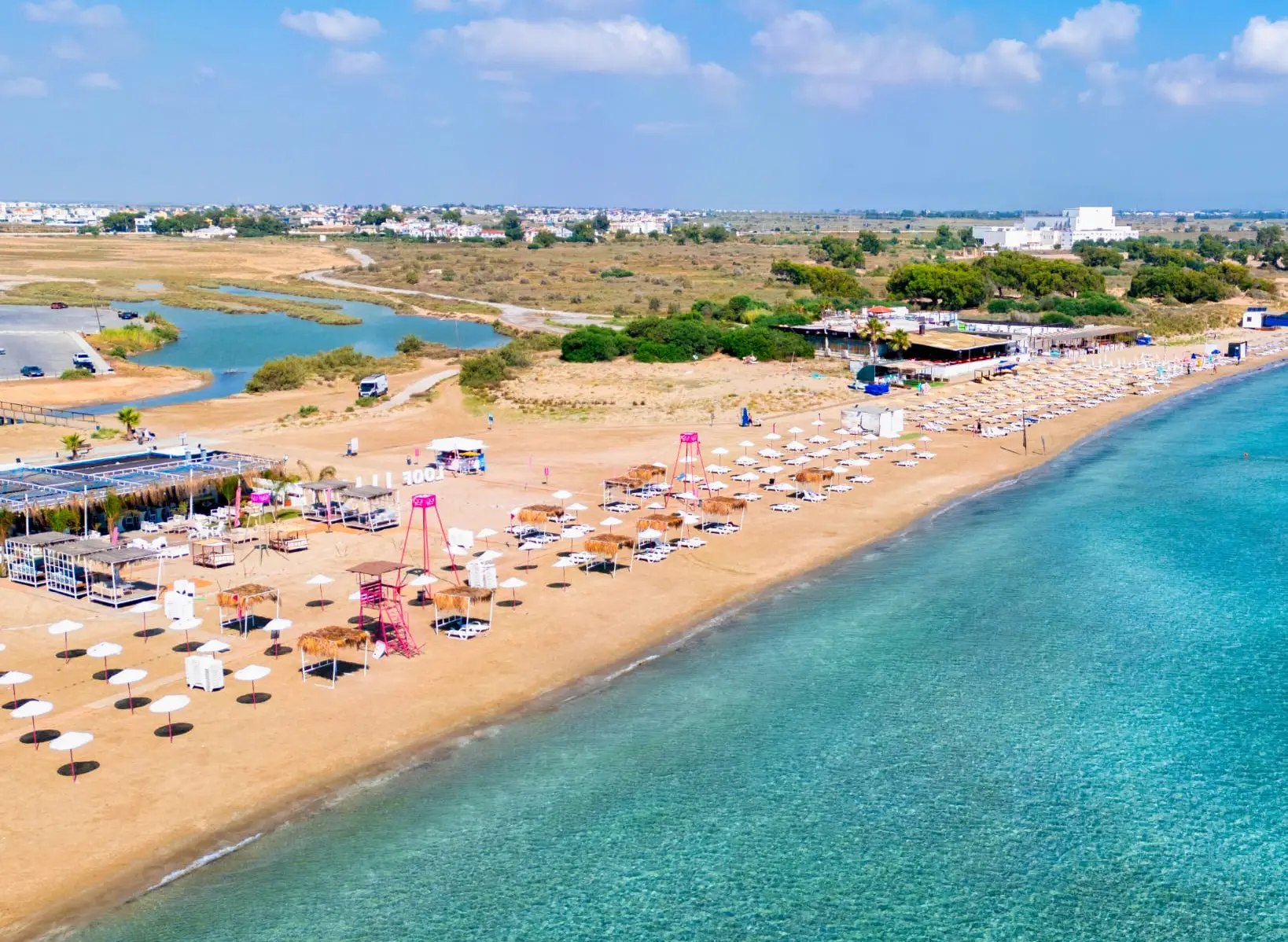 Kuzey Kıbrıs'ta turkuaz denize kıyısı olan bir plajın havadan çekilmiş görüntüsü; sıra sıra şezlong ve şemsiyeler, sazdan gölgelikler ve arka planda yeşillik ile kıyı yerleşimi görülüyor